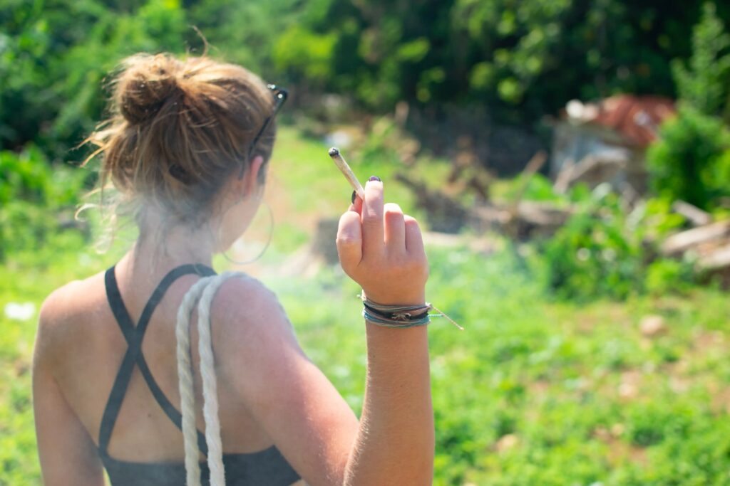A person with a bun hairstyle holds a lit cigarette outdoors in Winter Haven, Florida, with lush greenery in the background—perhaps taking a break after visiting a mobile smoke shop.