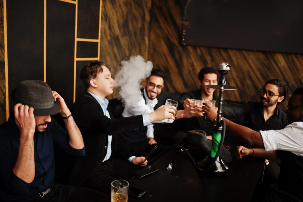 Five men sit around a table in a Winter Haven lounge, clinking glasses and smiling. One man exhales smoke as a hookah from a mobile smoke shop sits on the table with drinks and a phone.