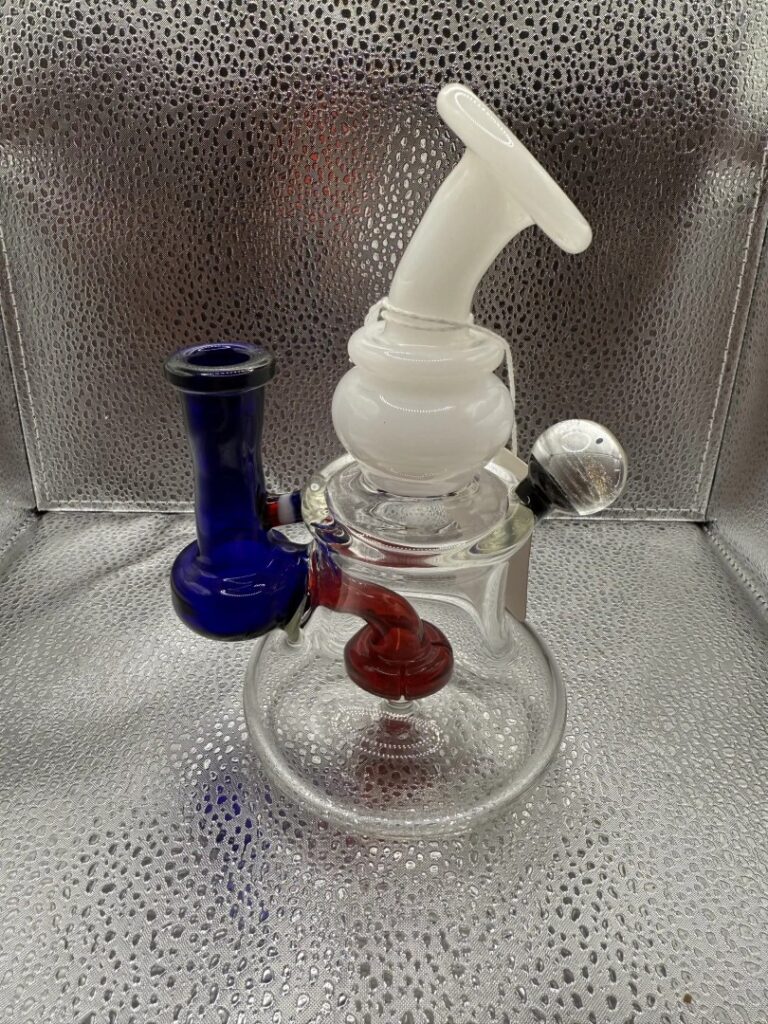 A glass water pipe with a white mouthpiece, purple and red accents, and a clear base, set against a textured metallic background—available from your Winter Haven mobile smoke shop in Florida.
