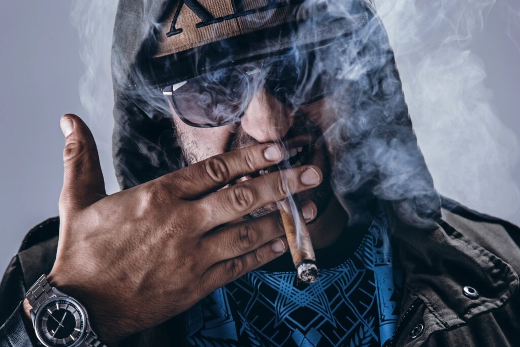 A person in a hooded jacket, sunglasses, and a cap holds a lit cigar near their mouth, surrounded by smoke—capturing the cool vibe of Winter Haven and the laid-back spirit of a Florida mobile smoke shop.