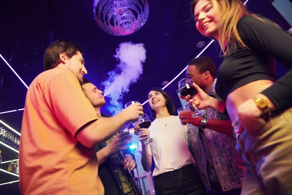 Five people stand in a Winter Haven nightclub holding drinks, smiling, and talking; neon lights from a mobile smoke shop and a chandelier shine overhead, with vapor swirling in the air.