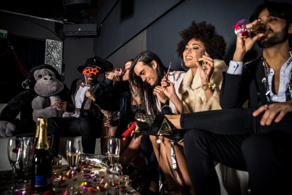 A group of people dressed in party attire sit on a couch with drinks and party favors, surrounded by confetti, a large stuffed gorilla, and mobile smoke shop accessories in Winter Haven, Florida.
