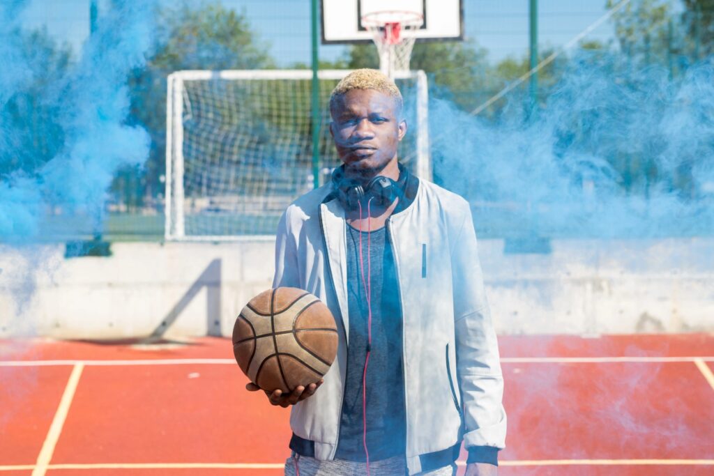 A person with headphones around their neck holds a basketball on an outdoor court in Winter Haven, Florida, surrounded by blue smoke. A basketball hoop and net are visible in the background.